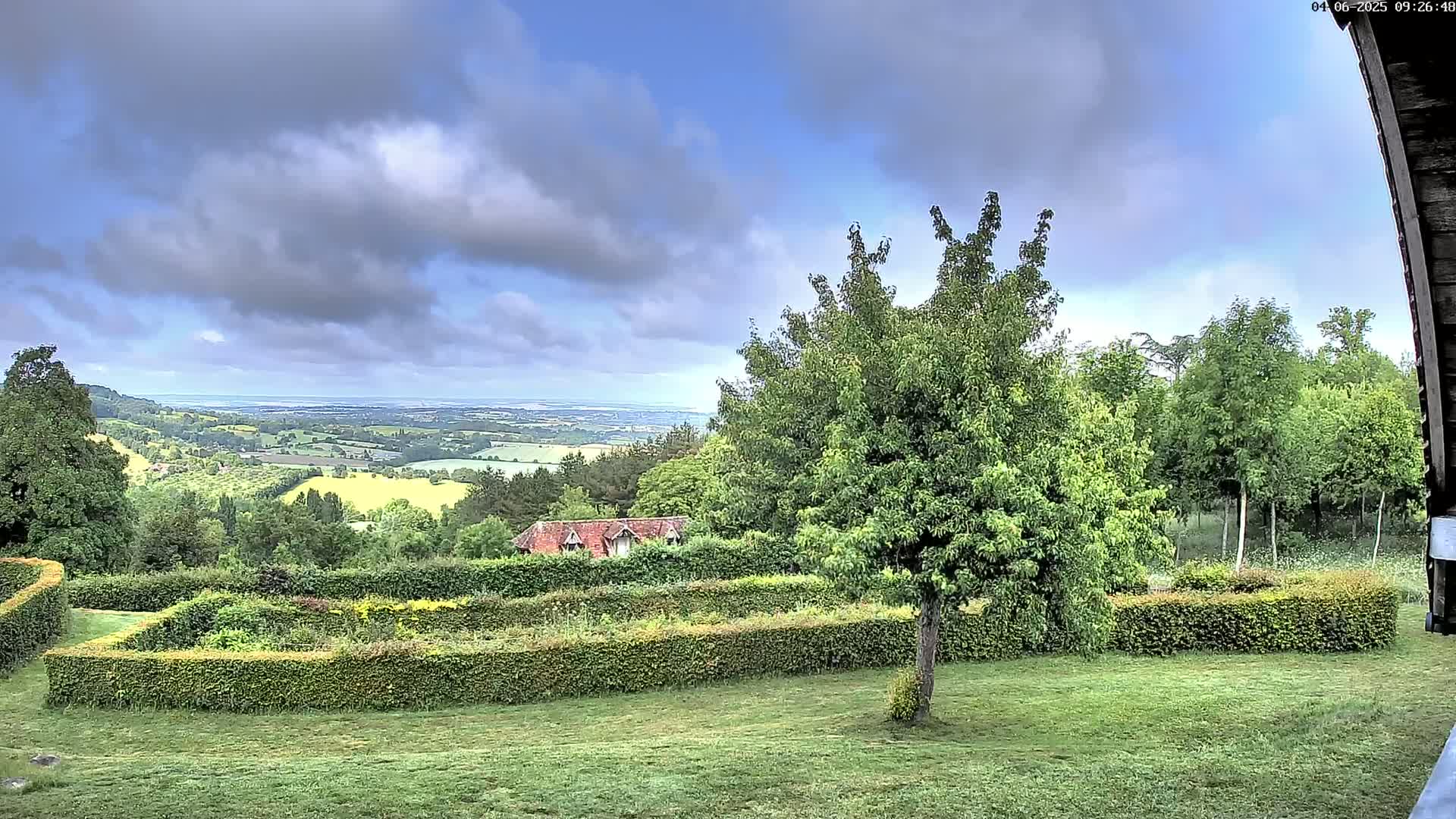 A partly cloudy day reveals a rolling green landscape with a distant body of water, viewed from a garden featuring manicured hedges and a tree.