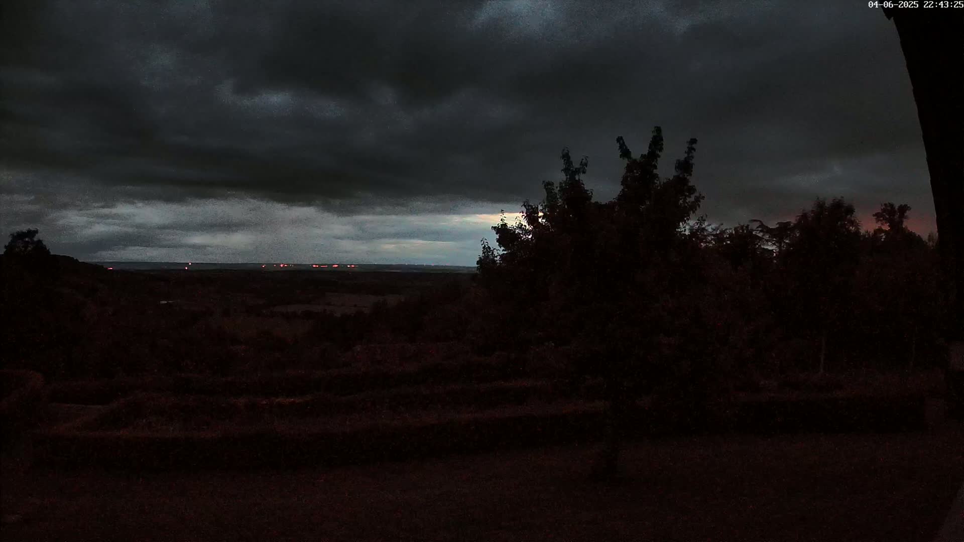 A dark, overcast sky hangs over a silhouetted landscape of trees and distant lights under the gloomy conditions.