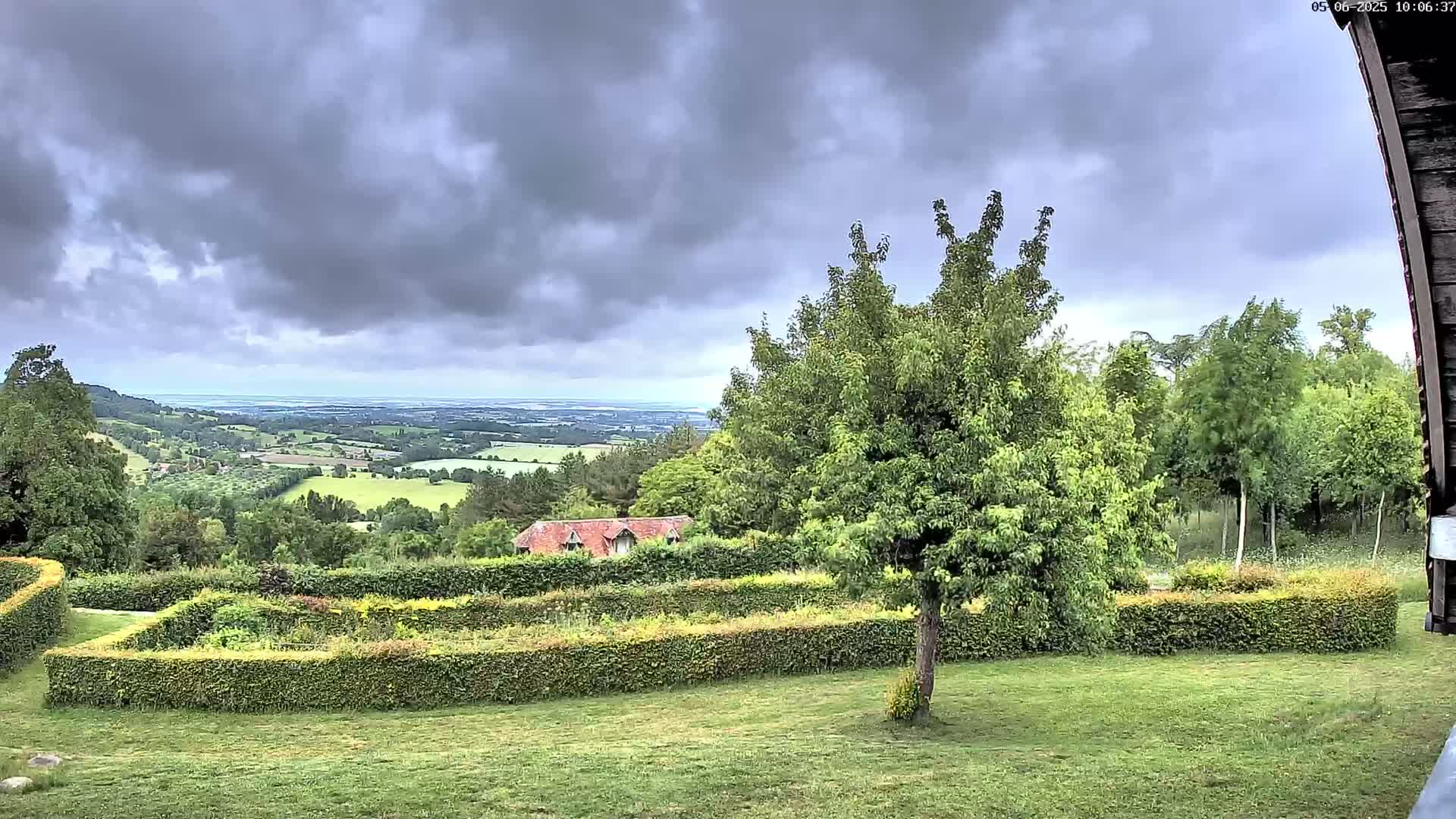A sprawling green landscape under a cloudy sky features a manicured garden with hedges, a small building, and a distant view of rolling hills and a body of water.