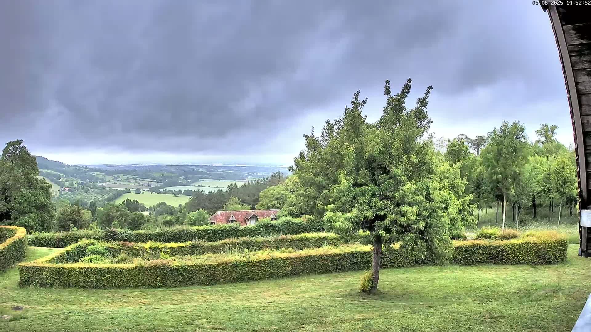 A verdant, hilly landscape under a cloudy sky is viewed from a garden containing a hedged enclosure and a single tree, with a small building visible in the mid-ground.