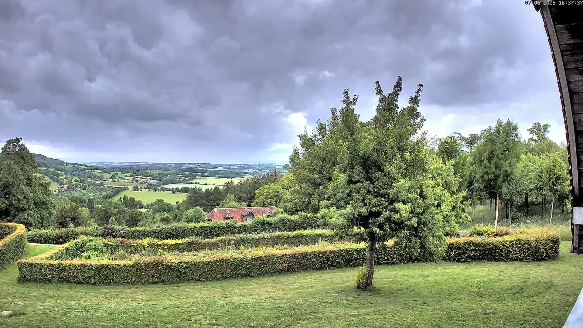 A verdant landscape featuring a neatly manicured garden with hedges, a tree, and a distant view of rolling hills under a cloudy sky.