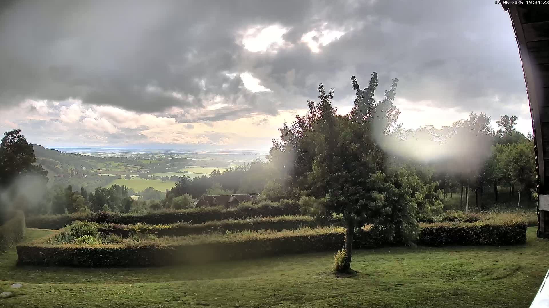 A hilly landscape with green fields and trees is visible under a partly cloudy sky, with some sunlight breaking through the clouds.