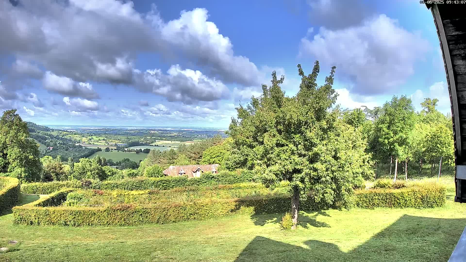 A partly cloudy day reveals a scenic view of a verdant valley, a distant body of water, and a meticulously landscaped garden with hedges.