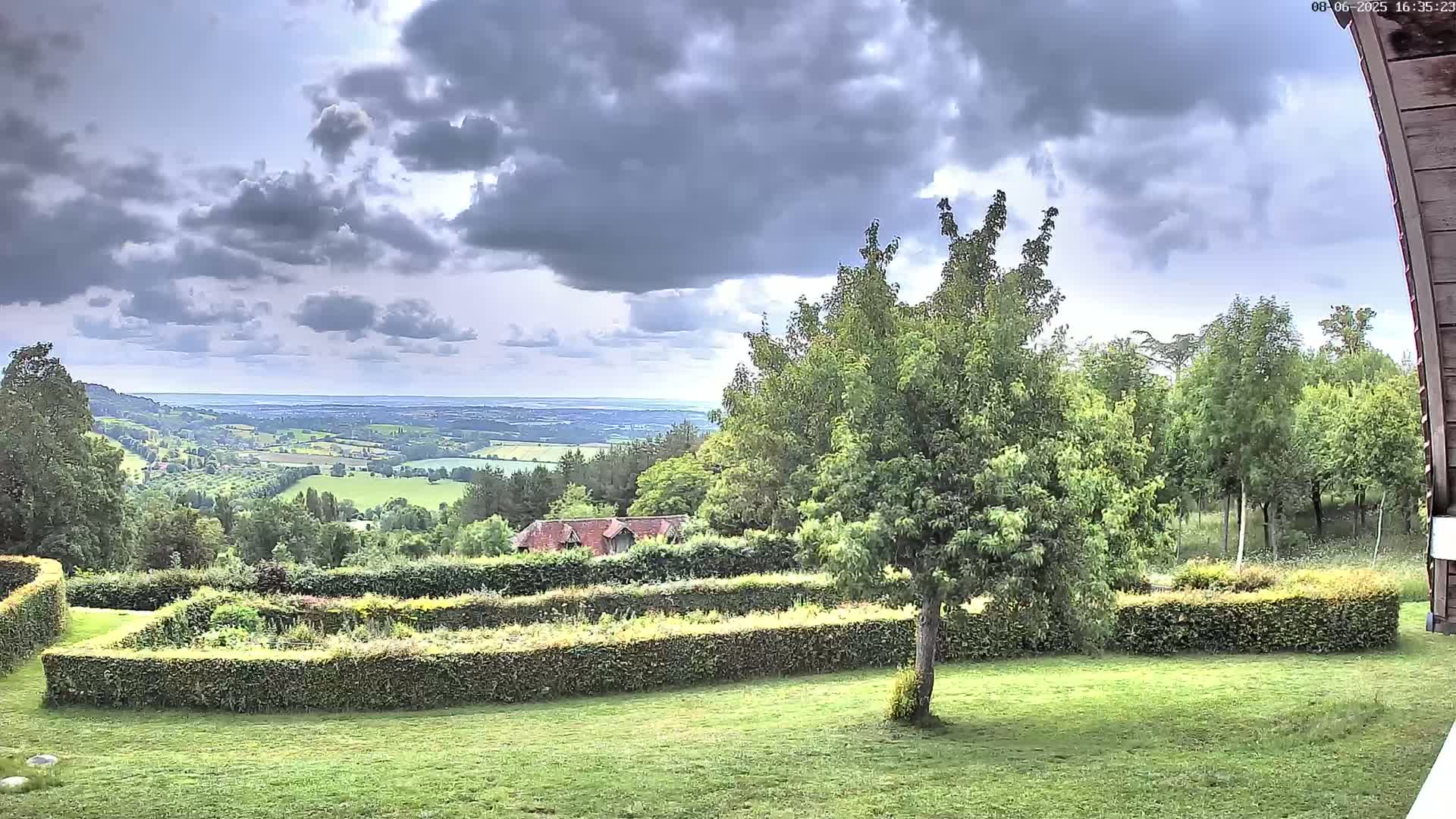 A verdant landscape with a neatly trimmed hedge encloses a garden, overlooking a sprawling valley under a partly cloudy sky.