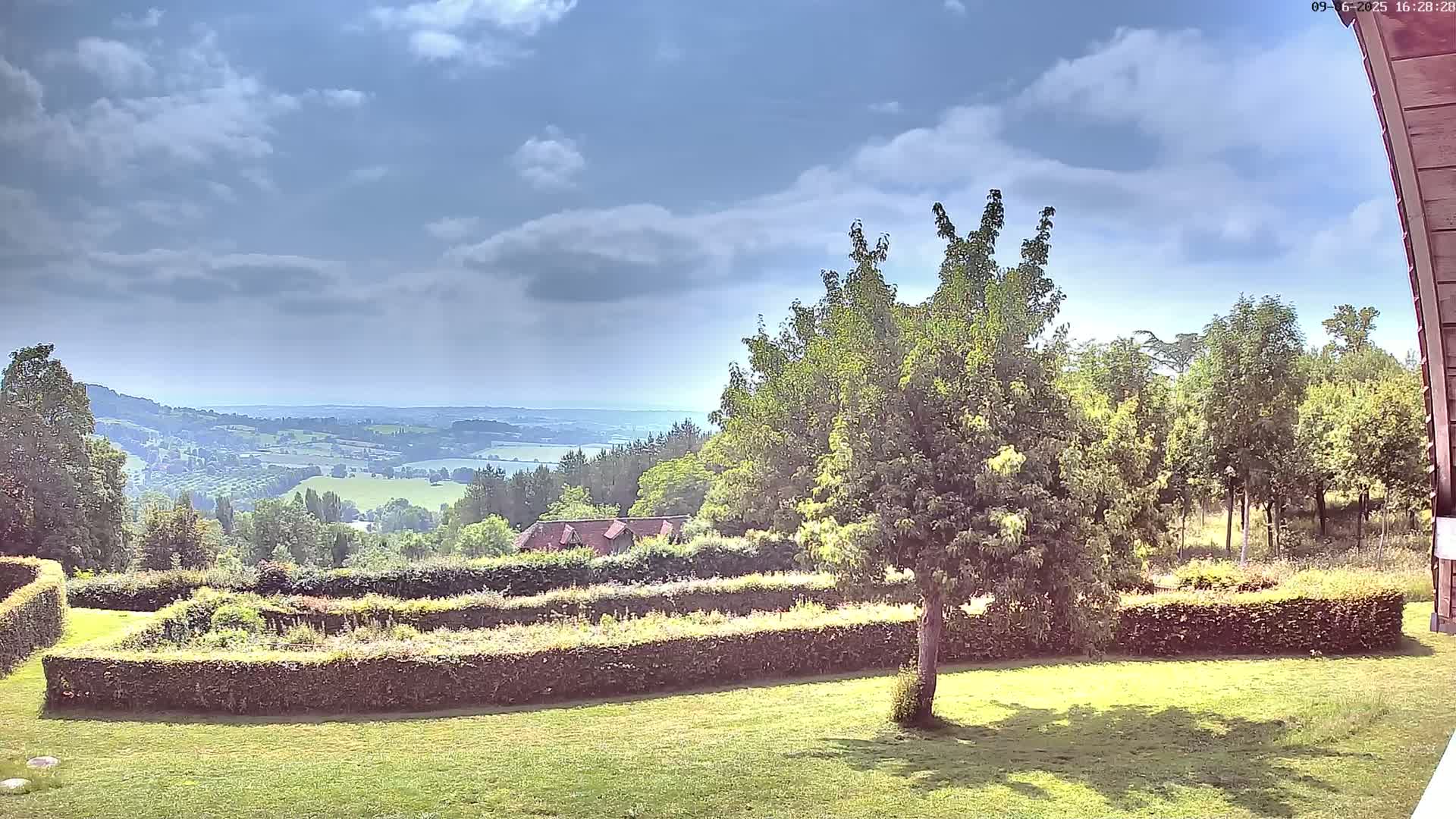 A partly sunny day reveals a verdant landscape featuring a manicured lawn, hedges forming geometric patterns, and a distant view of rolling hills and a body of water under a partly cloudy sky.