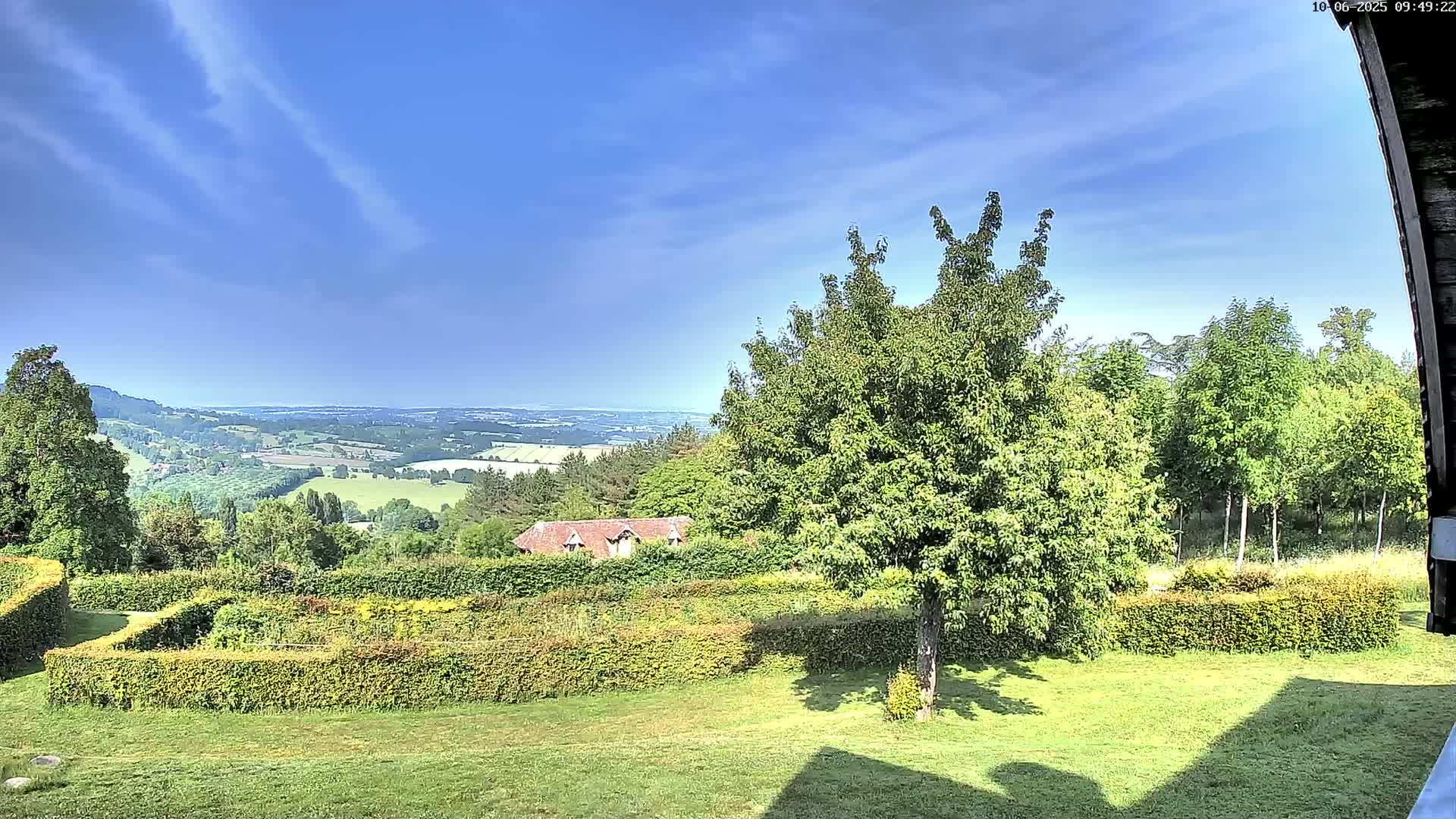 A verdant, manicured lawn with neatly trimmed hedges overlooks a sprawling valley under a clear, bright blue sky with wispy clouds.