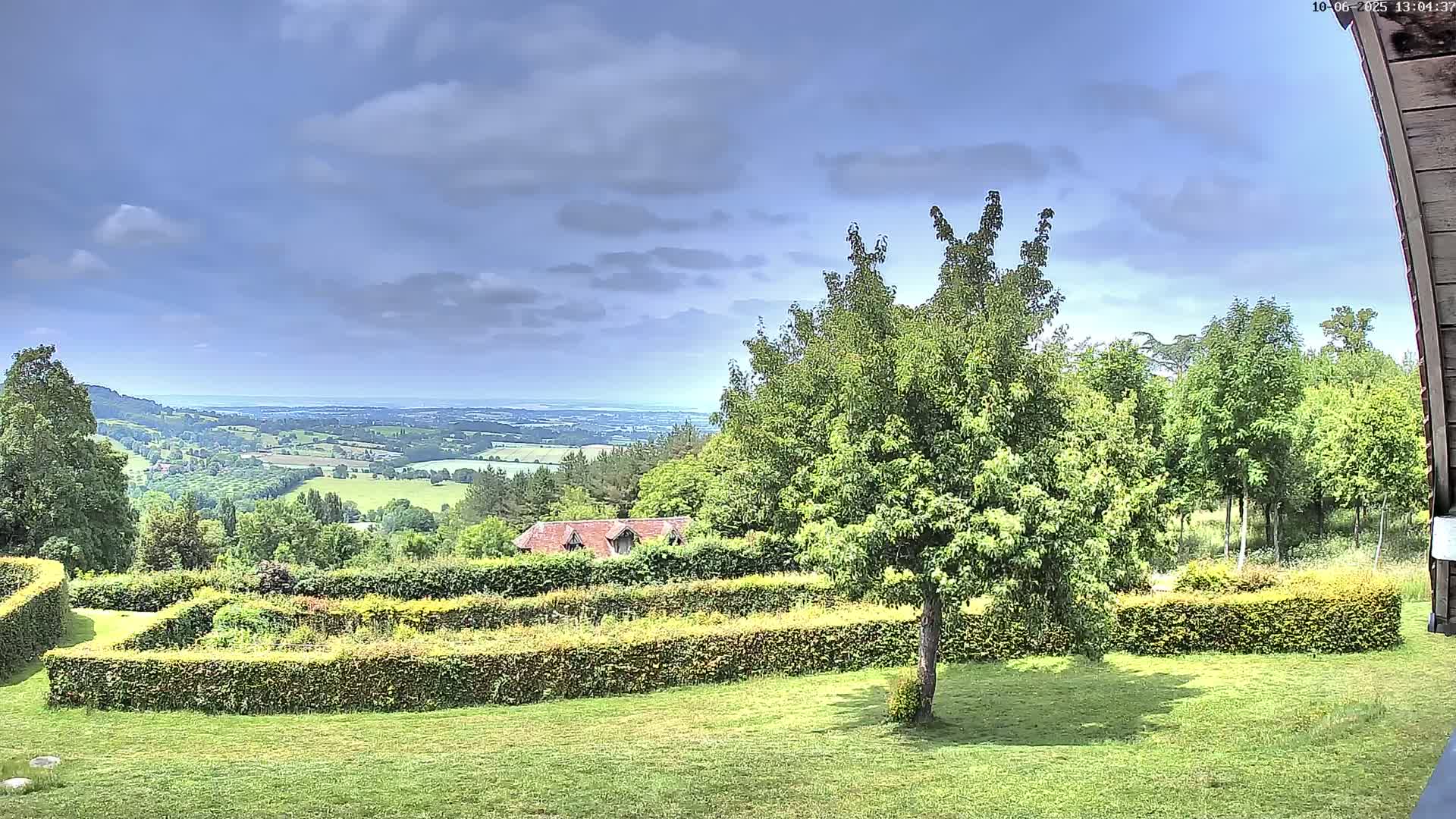 A partly cloudy sky overlooks a verdant landscape featuring a manicured garden with hedges, a tree, and a distant view of rolling hills and a body of water.