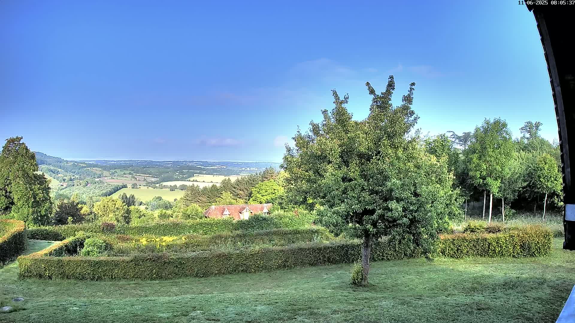 A verdant landscape under a clear blue sky shows a manicured garden with hedges, a house nestled in the trees, and a distant view of rolling hills.