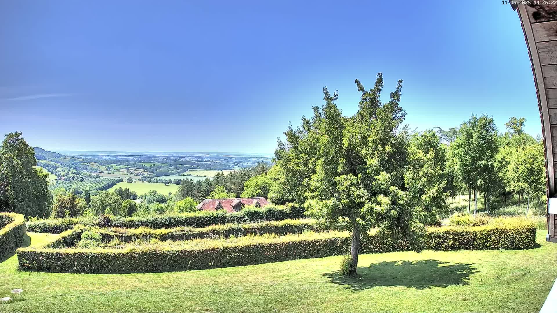 A lush green lawn with neatly trimmed hedges overlooks a valley and distant body of water under a clear blue sky.