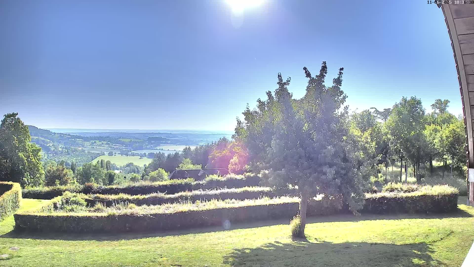 A sunny day reveals a verdant landscape with neatly trimmed hedges, a tree in the foreground, and a distant view of rolling hills and a body of water under a clear blue sky.