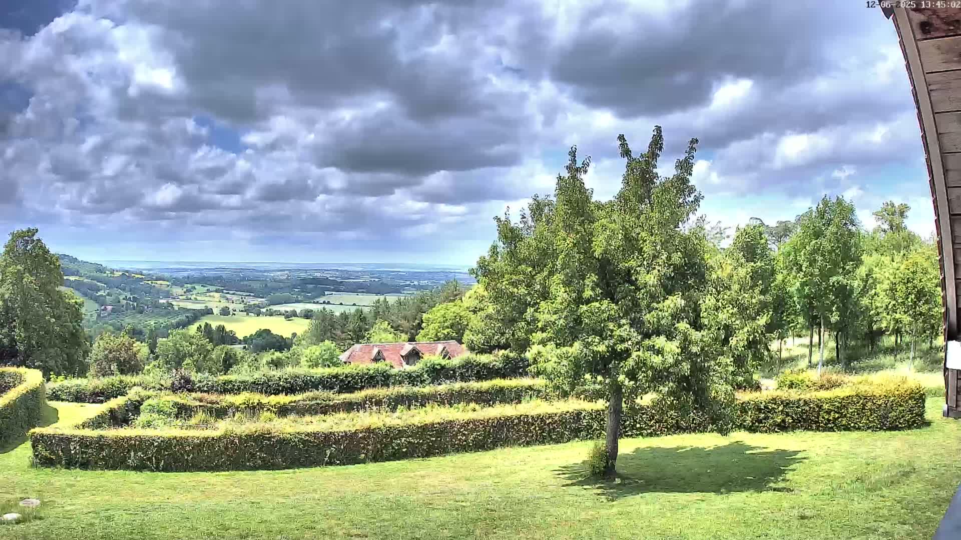 A partly cloudy day reveals a sprawling green landscape, featuring manicured hedges, a lone tree, and a distant view of a valley stretching to a body of water under a partly cloudy sky.