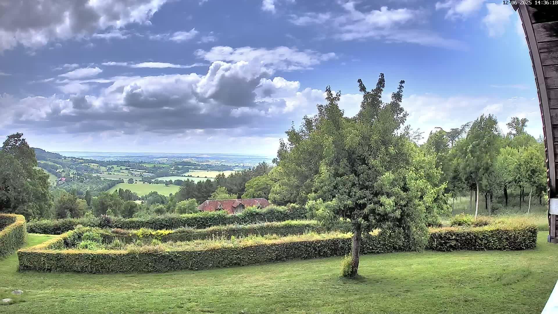 A partly cloudy sky overlooks a verdant landscape featuring a manicured garden, a small building, and a distant valley extending to a hazy horizon.