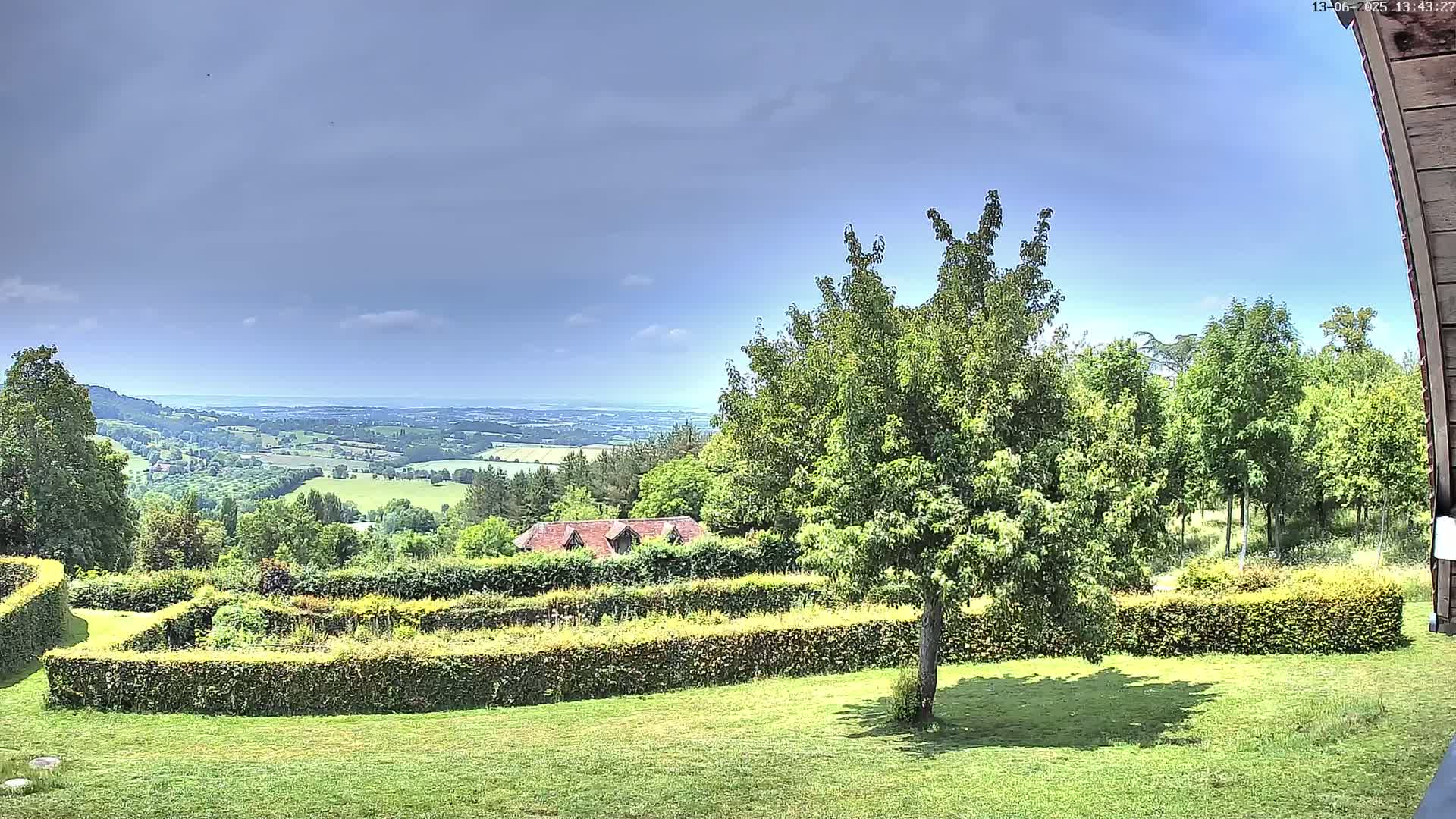 A partly cloudy sky overlooks a grassy lawn with a manicured hedge surrounding a garden, a tree in the foreground, and a distant view of rolling green hills and a body of water.