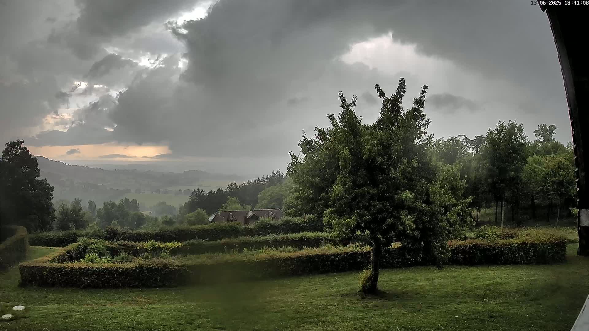 A rain-swept landscape shows a green lawn, hedged garden, and trees under a dark, cloudy sky with a hint of sunset in the distance.