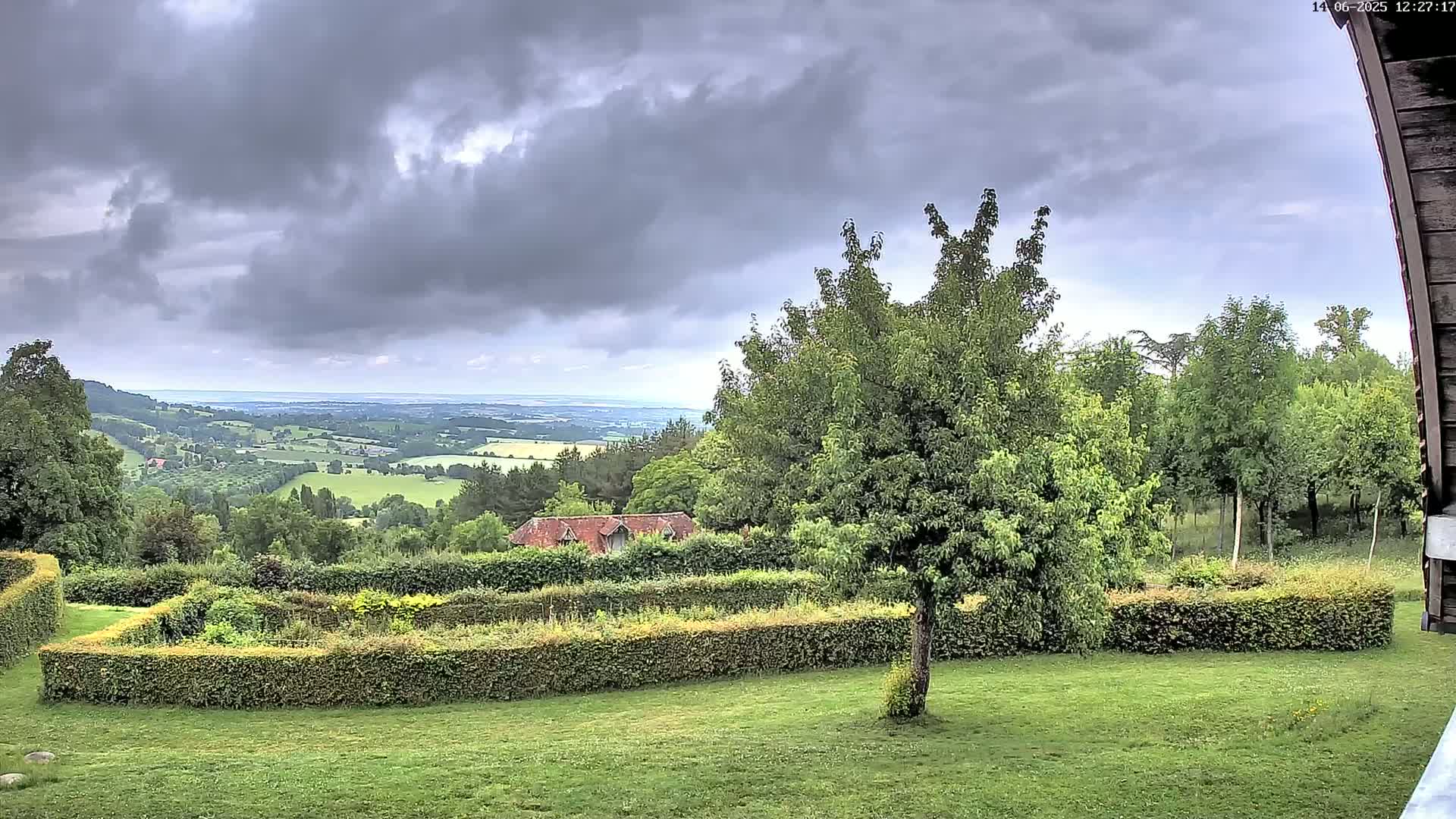 A verdant landscape featuring a manicured garden with hedges, a small building, and numerous trees under a cloudy sky.