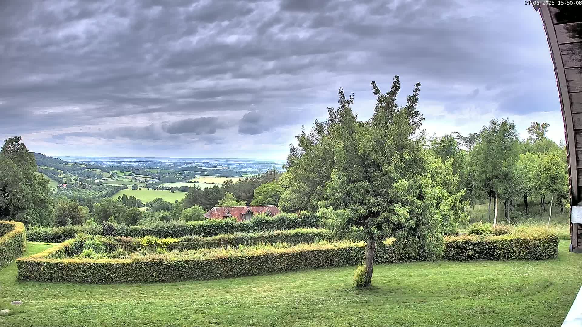 A grassy yard with manicured hedges overlooks a rolling green valley under a cloudy sky.