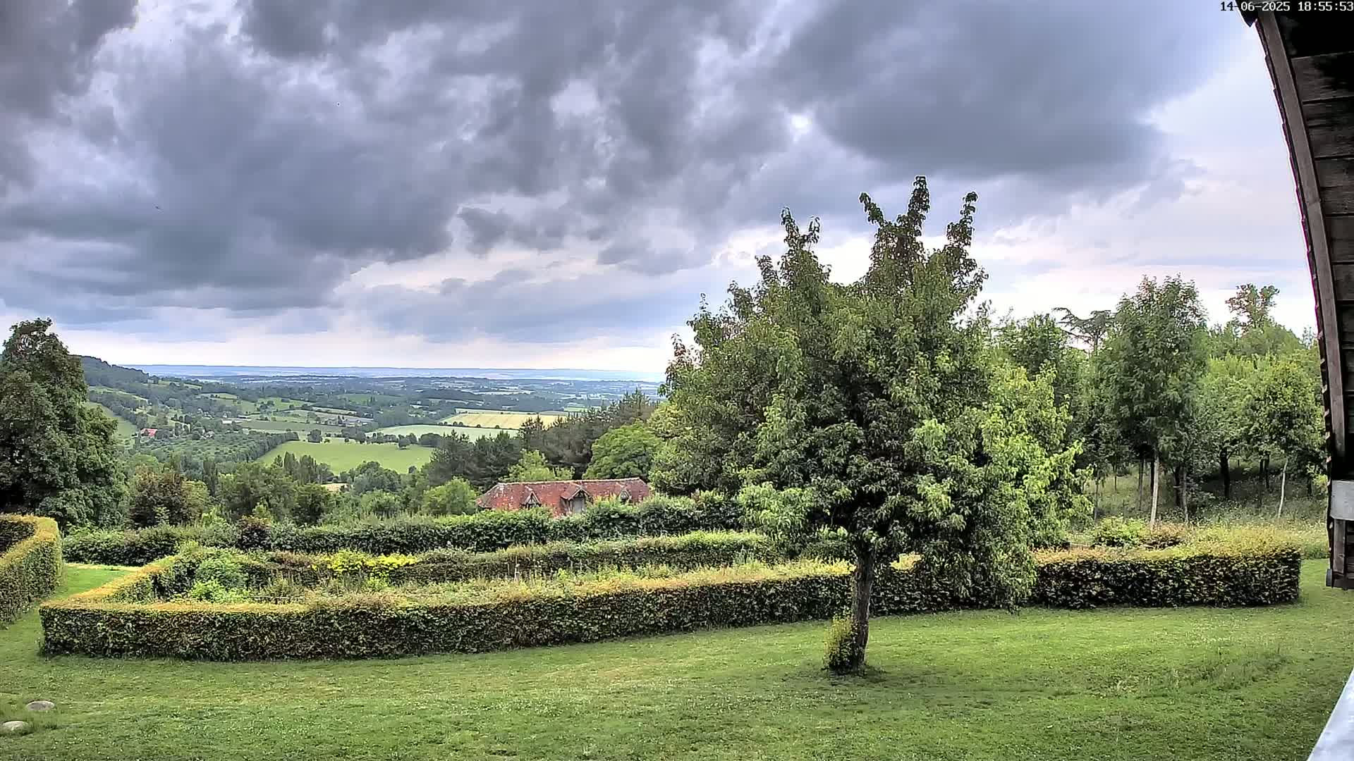 A grassy yard with manicured hedges overlooks a rolling green valley under a cloudy sky.