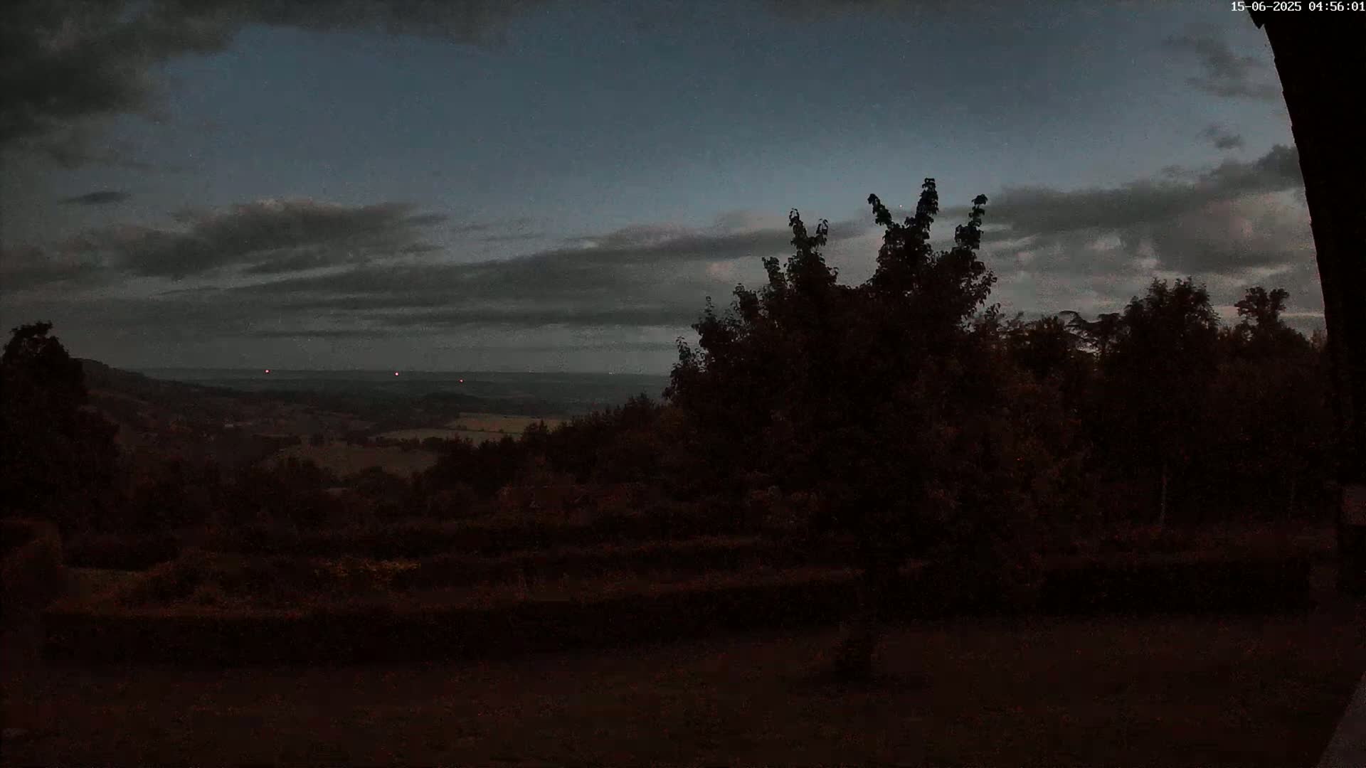 A dark, overcast sky hangs over a hilly landscape, partially obscured by silhouetted trees and shrubs at dusk.