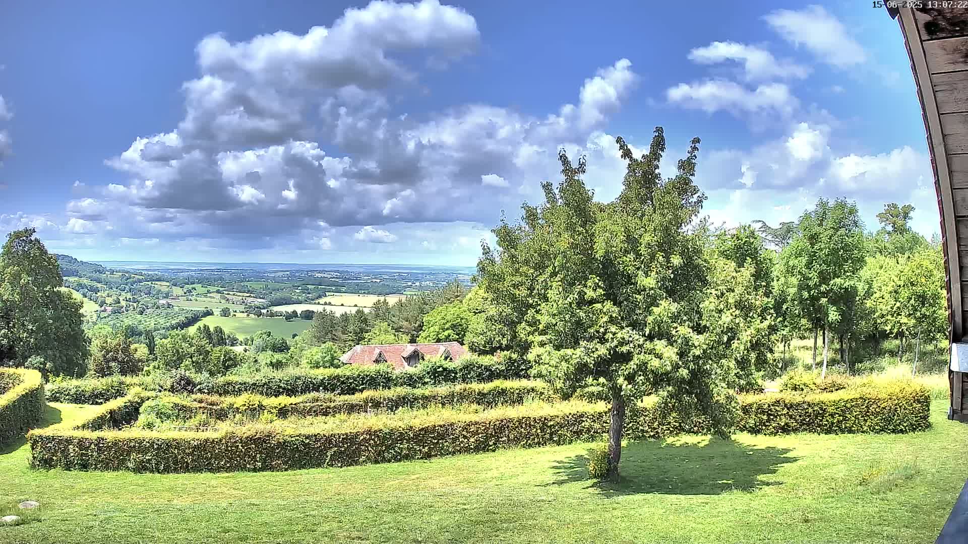 A partly sunny view from a garden shows a rolling green landscape extending to a distant body of water under a partly cloudy sky.