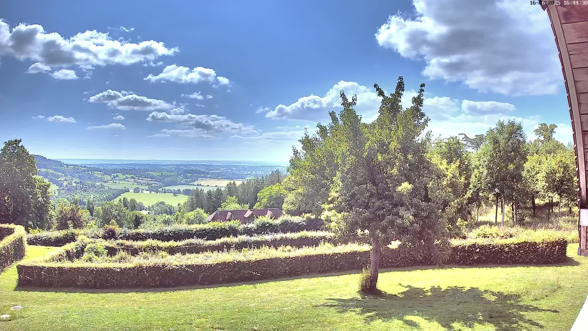 A sunny day reveals a sprawling green landscape, featuring manicured hedges, lush trees, and a distant view of rolling hills under a partly cloudy sky.