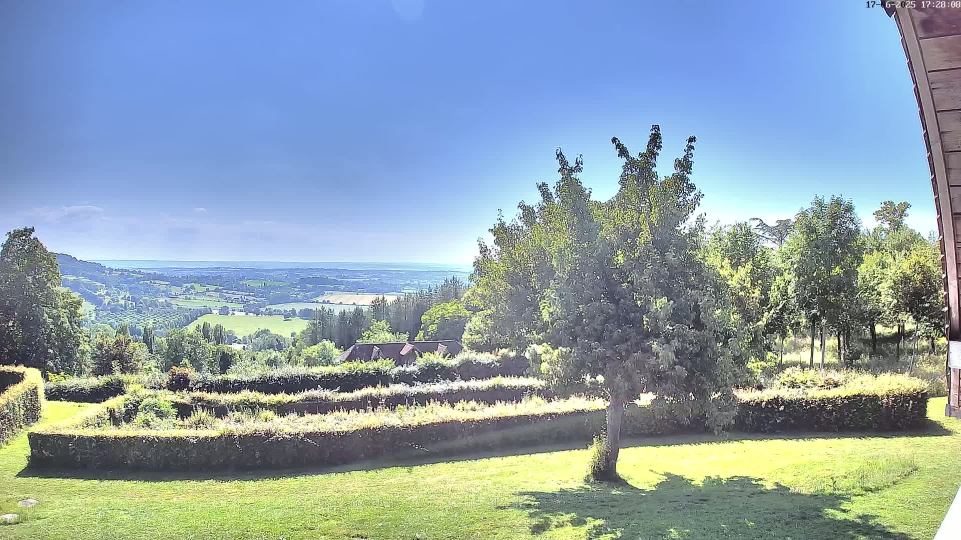 A sunny day reveals a verdant landscape featuring a manicured garden with hedges, a large tree, and a distant view of rolling hills and a body of water under a clear blue sky.