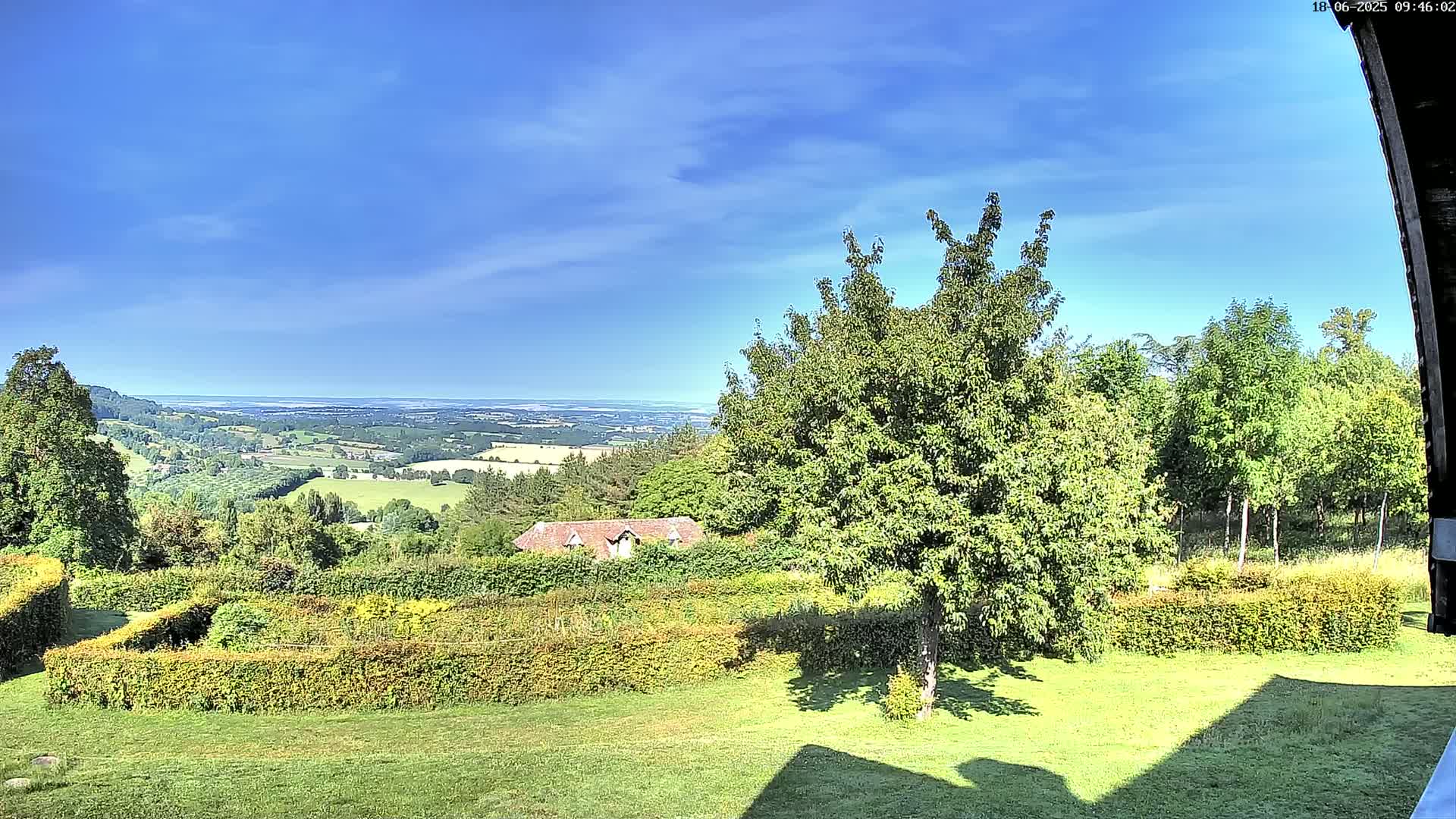 A hilly landscape with a neatly trimmed hedge and a small house is visible under a clear blue sky, overlooking a distant, flat expanse of land.