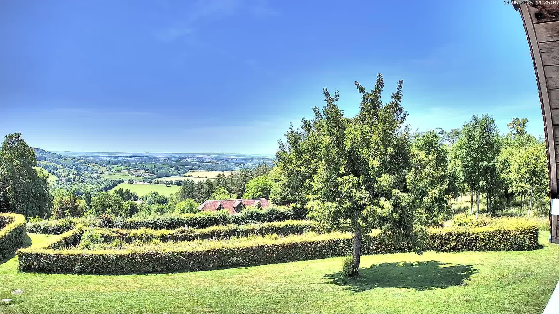 A sunny day reveals a sprawling green landscape with a meticulously manicured garden featuring hedges, a distant view of rolling hills and a body of water, and a small building nestled amongst the trees.
