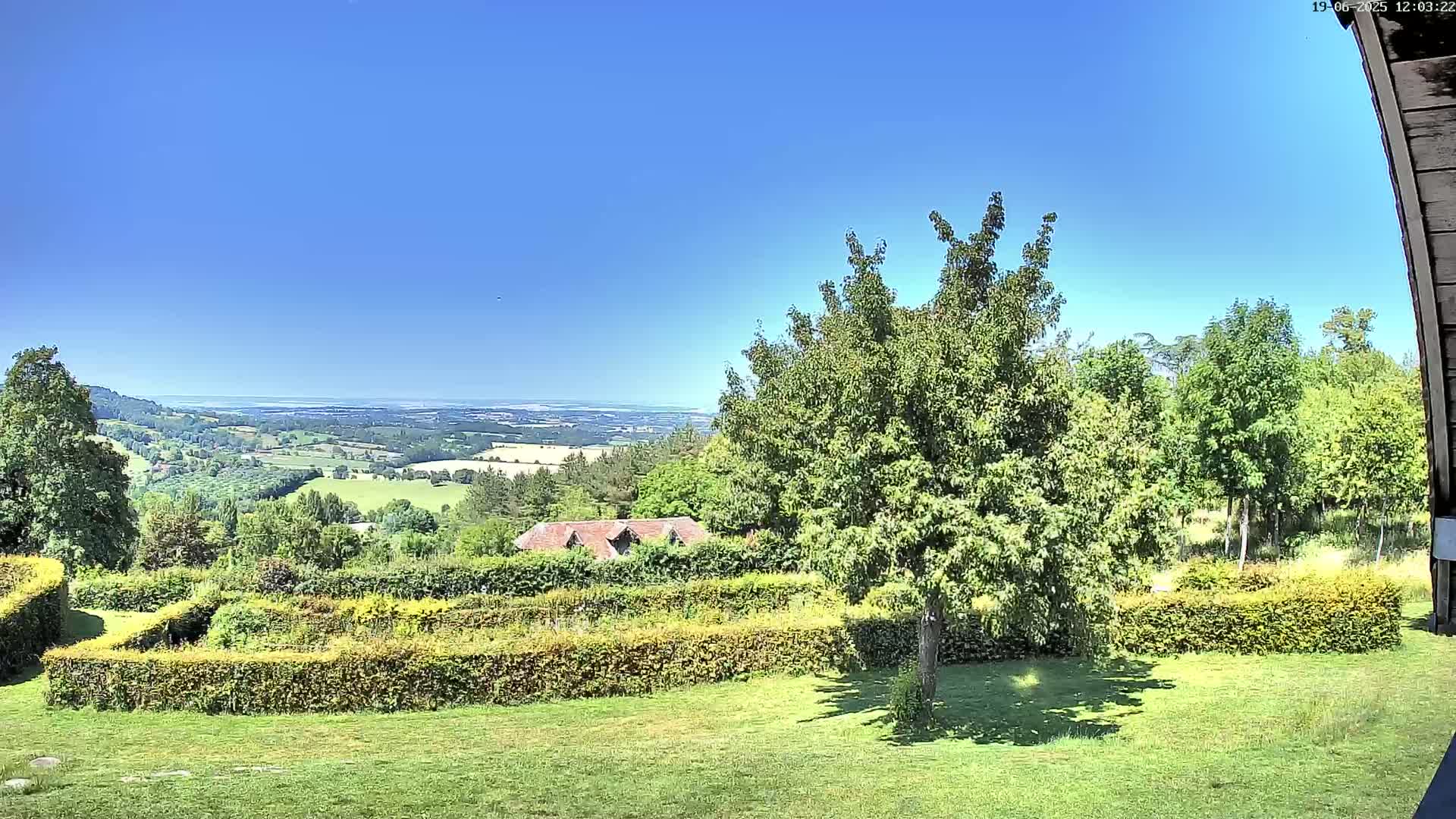 A verdant landscape under a clear blue sky shows a manicured lawn, neatly trimmed hedges, and a distant view of rolling hills and a body of water.