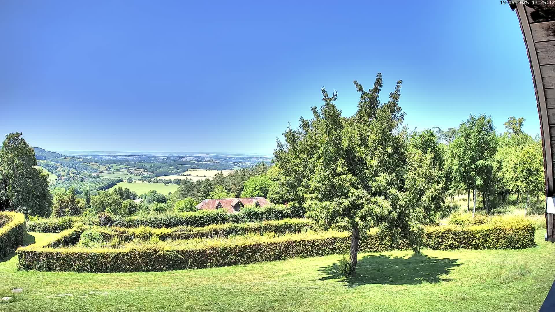A lush green lawn with a neatly trimmed hedge encloses a garden overlooking a vast, sunny valley and distant hills under a clear blue sky.