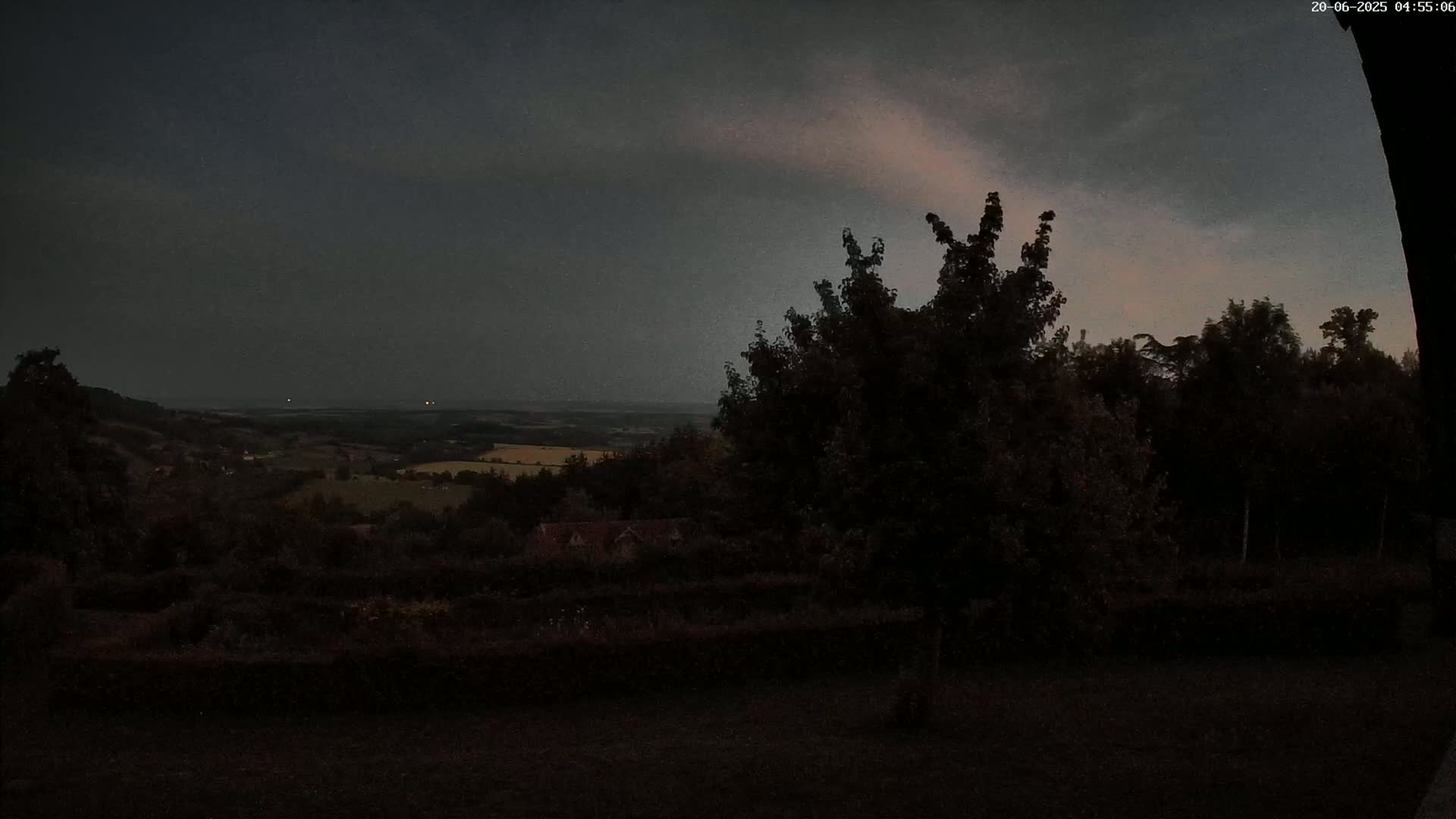 A dark, overcast sky hangs over a hilly landscape of dark green trees and fields, visible from a vantage point slightly elevated amongst more trees.