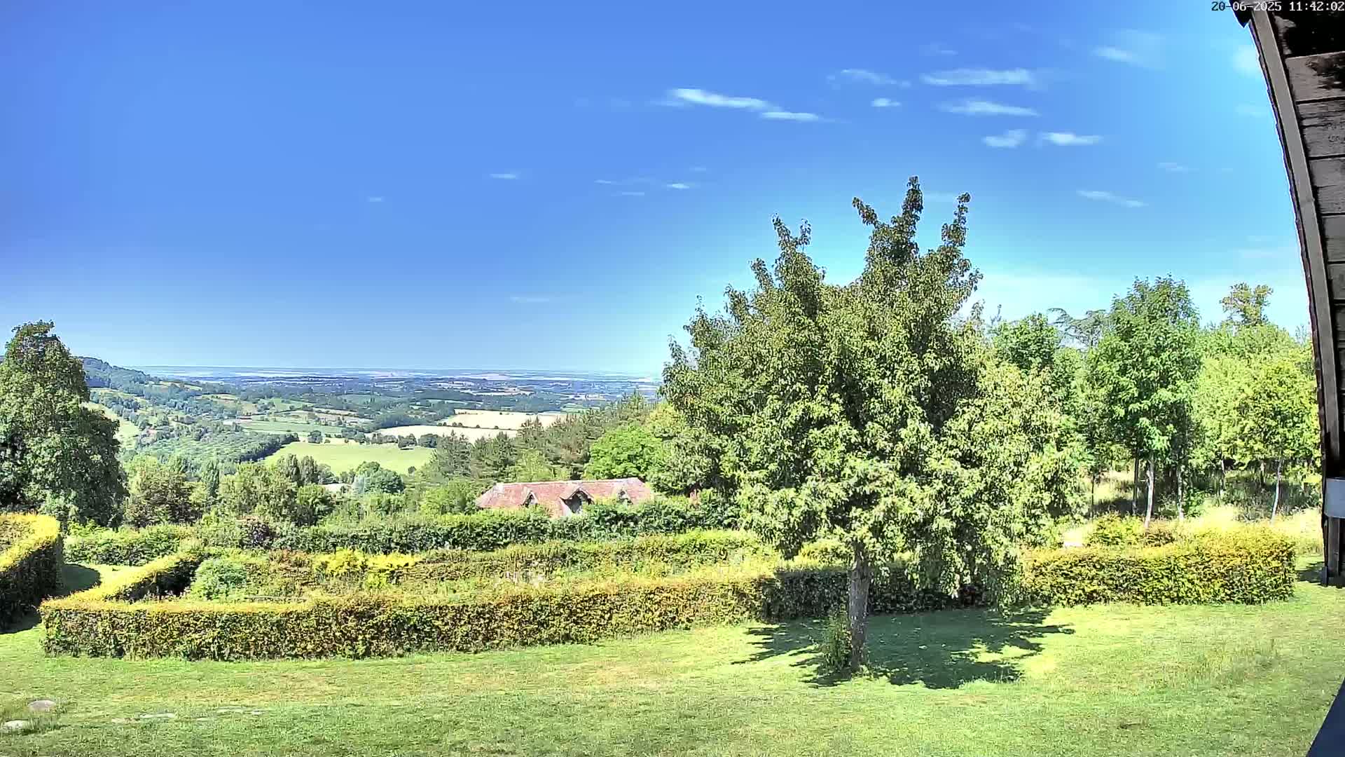 A sunny day reveals a verdant landscape, featuring a manicured lawn, neatly trimmed hedges, a tree in the foreground, and a distant view of rolling hills and a clear blue sky.