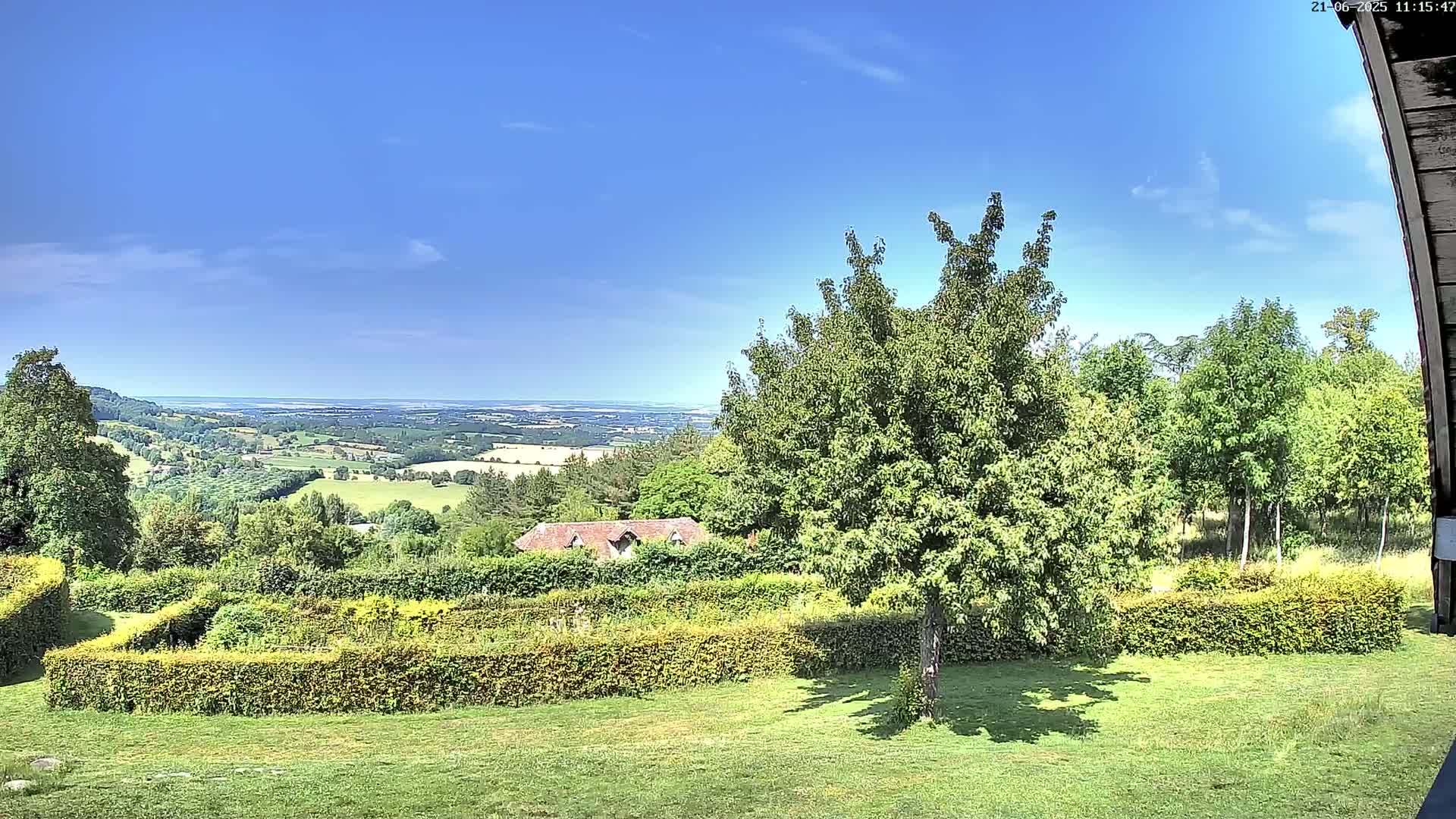 A sprawling green landscape with neatly trimmed hedges, a small house, and various trees under a mostly clear, sunny sky overlooks a distant valley.