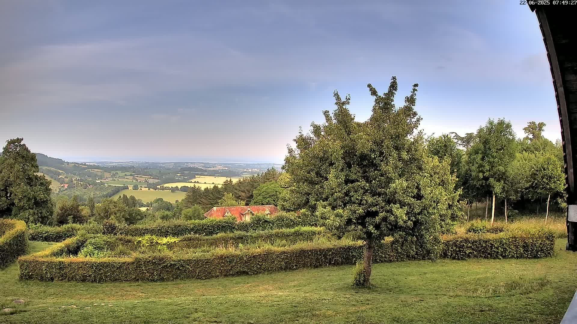 A partly cloudy sky overlooks a verdant landscape featuring a neatly trimmed garden, a small building, and a distant view of rolling hills and a body of water.