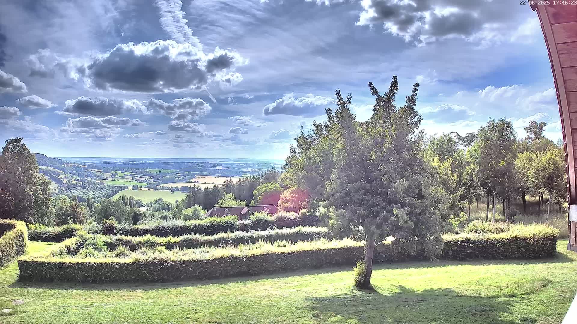 A partly cloudy sky overlooks a verdant landscape featuring a manicured garden, trees, and a distant view of rolling hills and fields.