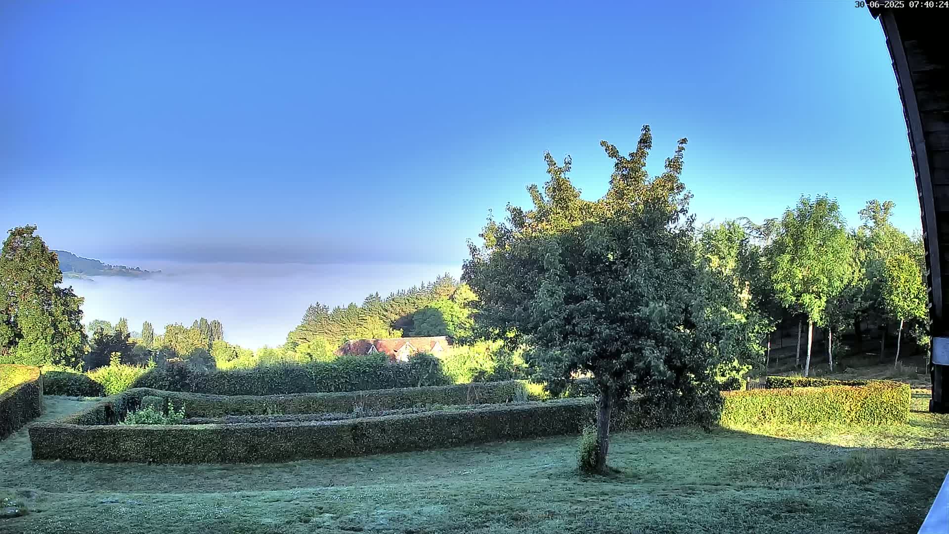 A neatly manicured garden with a low hedge enclosure overlooks a valley shrouded in morning mist under a clear blue sky.