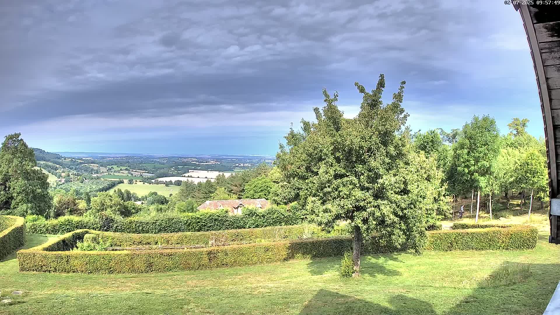 A verdant landscape under a mostly cloudy sky reveals a distant ocean view and a manicured garden with hedges.