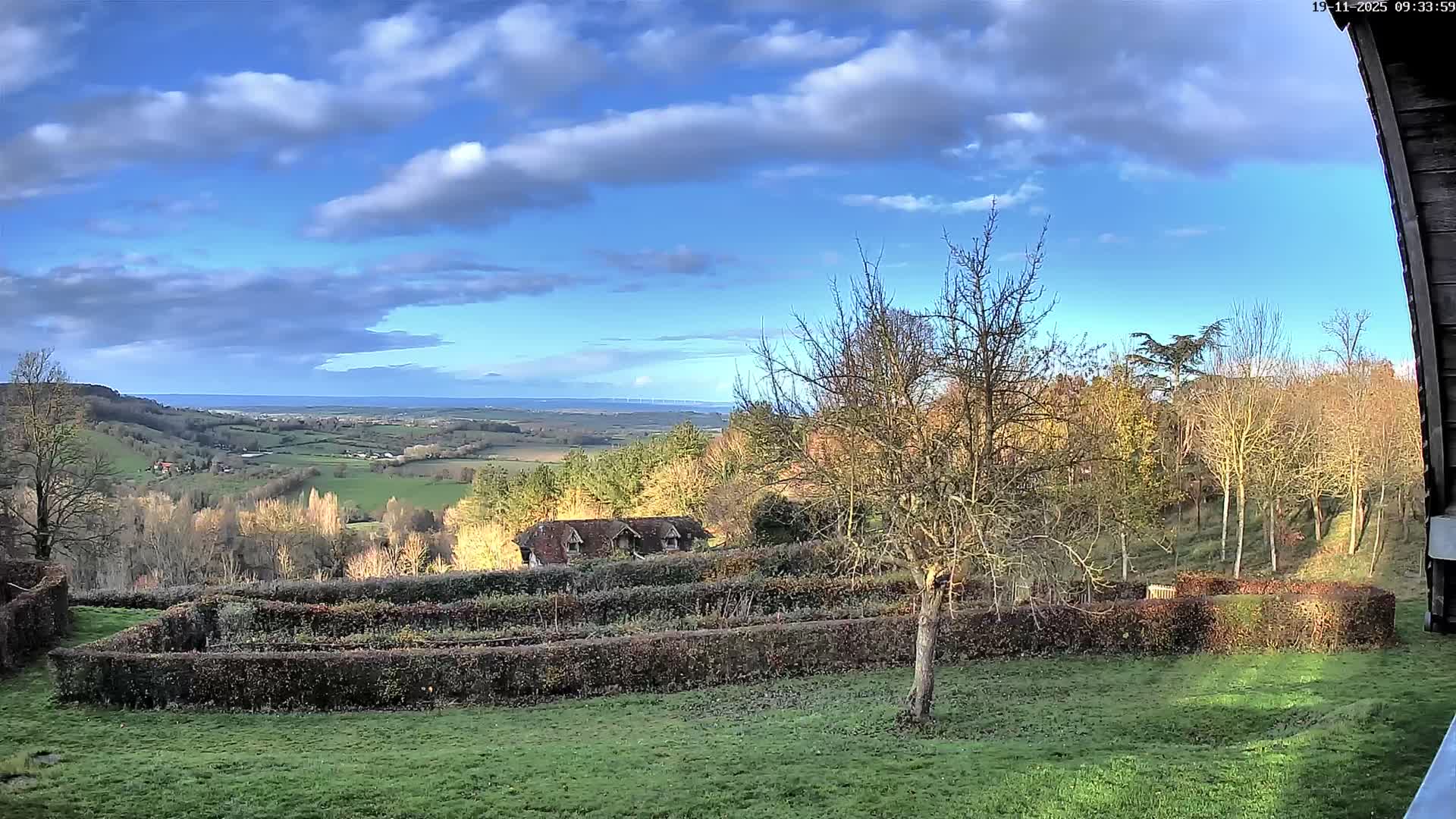 A bright, partly cloudy day illuminates a broad rural landscape, showcasing a grassy foreground with terraced hedges and a bare tree, leading to rolling green hills, a small building with dormers, and a distant body of water with a bridge on the horizon.