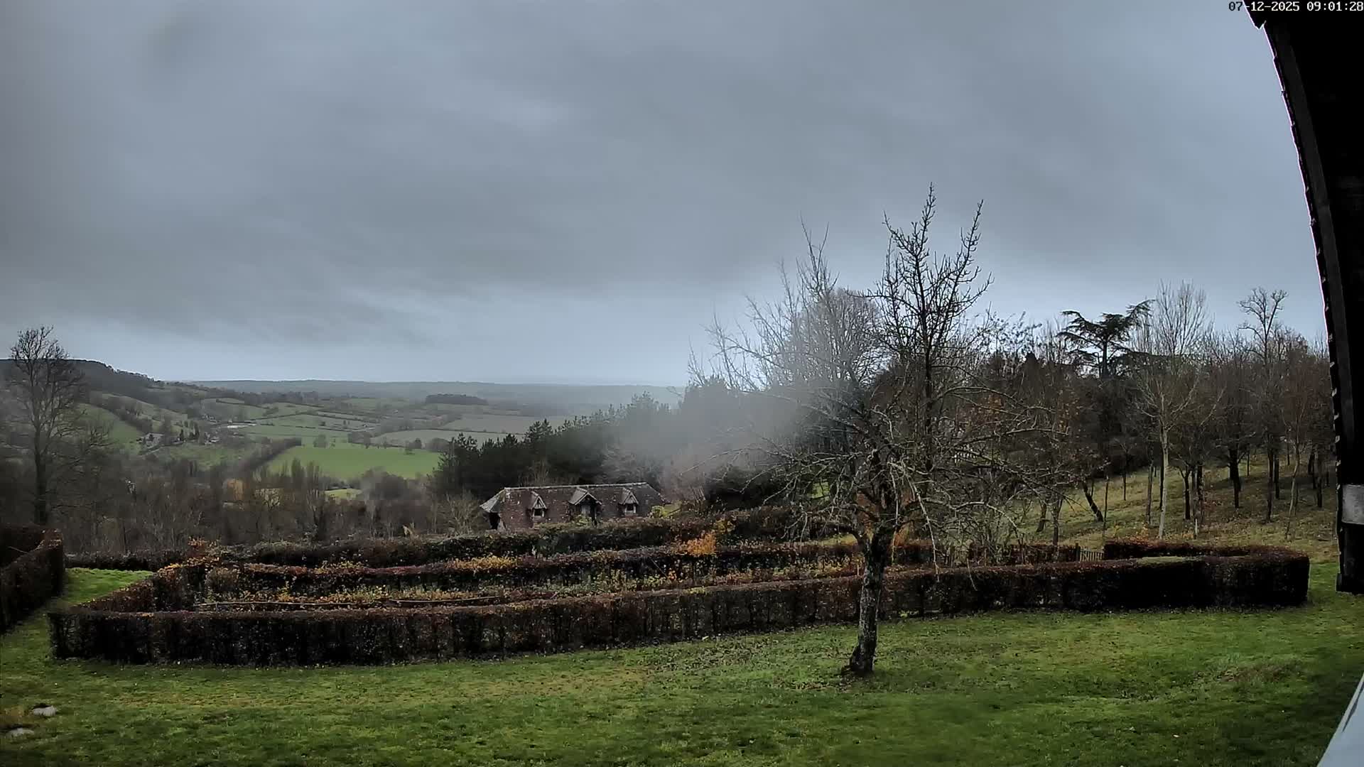 Under an overcast and misty sky, a green lawn and a dark hedgerow garden lead the eye to a partially obscured house nestled among rolling green and wooded hills stretching to the horizon.
