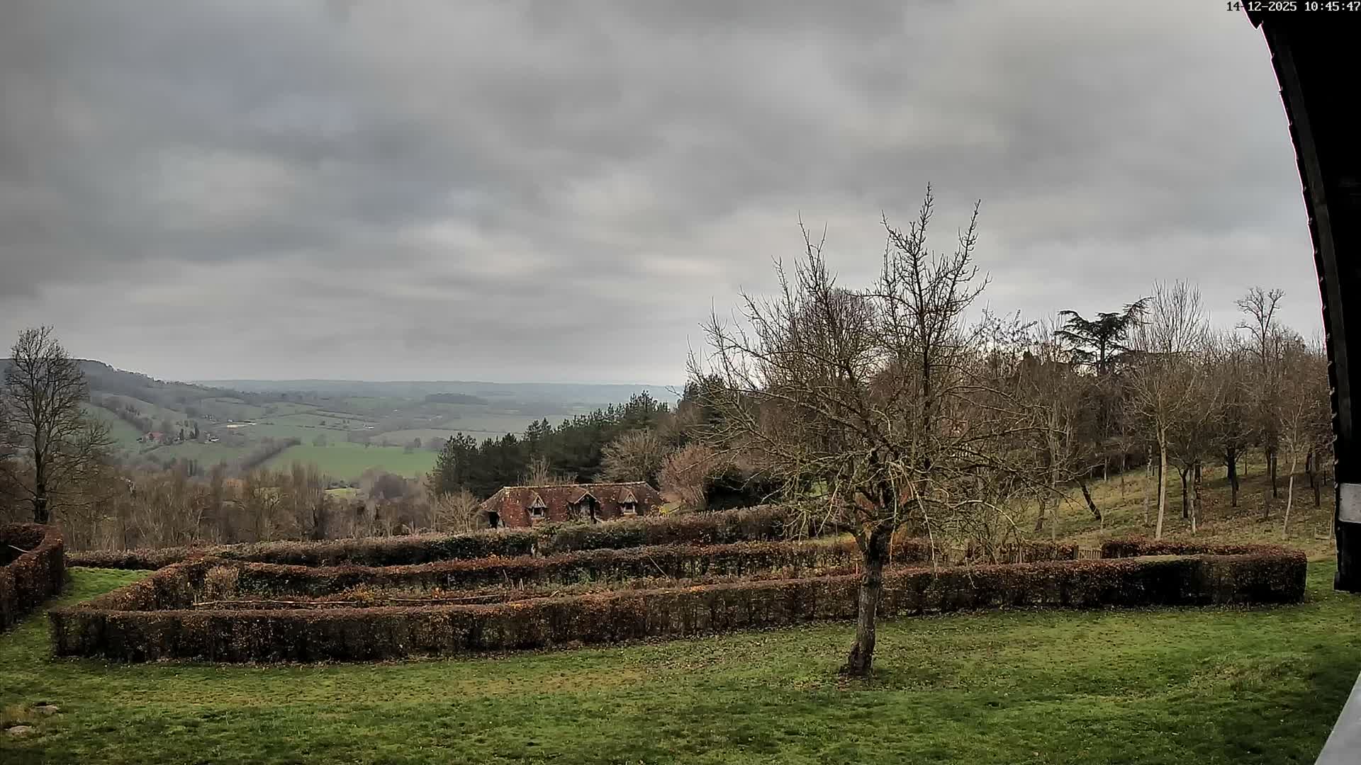 Under an overcast and misty sky, a green lawn and a dark hedgerow garden lead the eye to a partially obscured house nestled among rolling green and wooded hills stretching to the horizon.