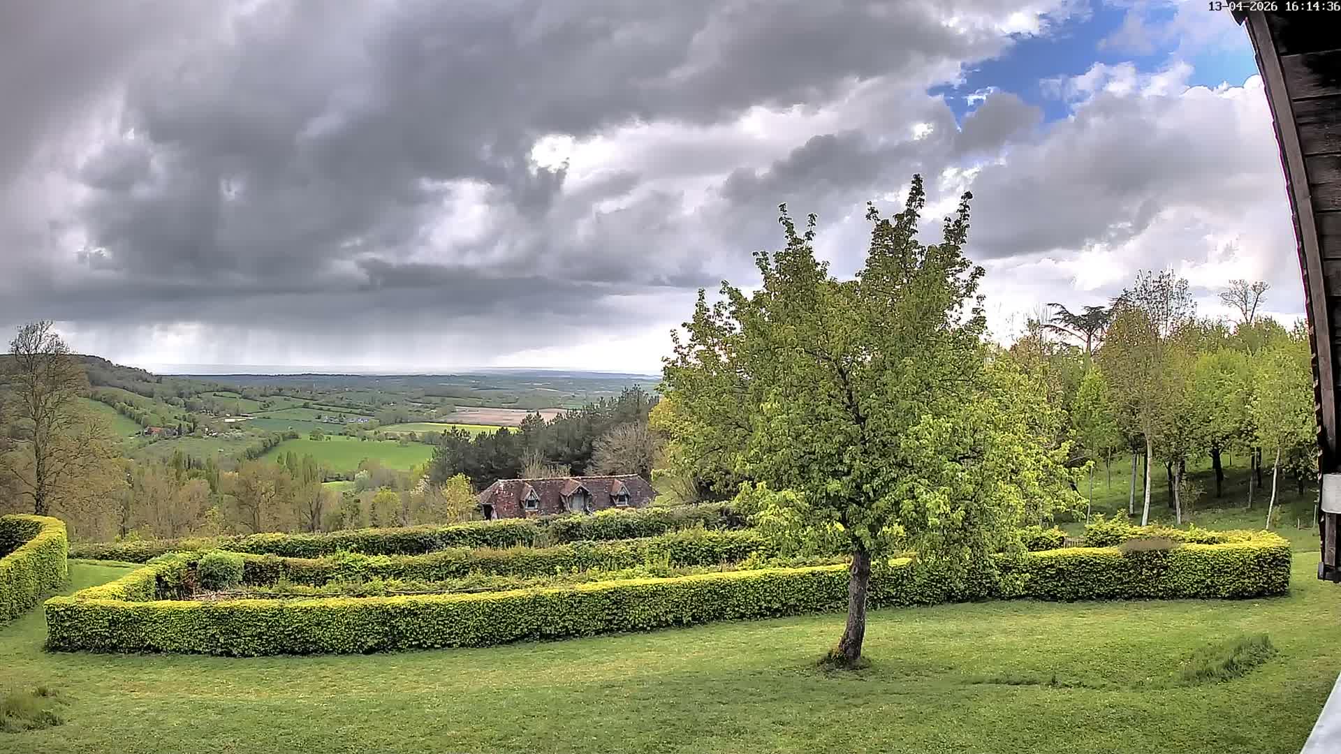 Under an overcast and misty sky, a green lawn and a dark hedgerow garden lead the eye to a partially obscured house nestled among rolling green and wooded hills stretching to the horizon.