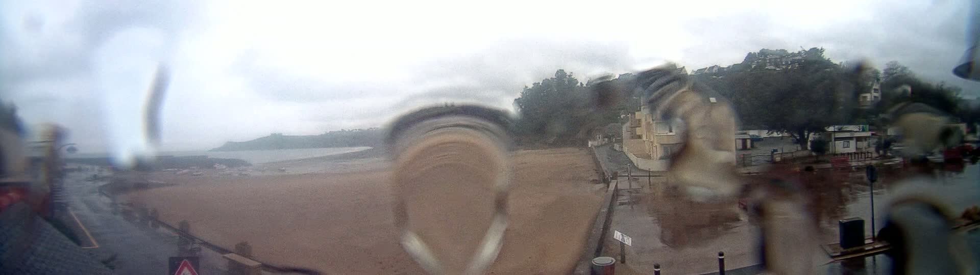 A rain-streaked window shows a view of a beach, buildings, and trees on an overcast day.