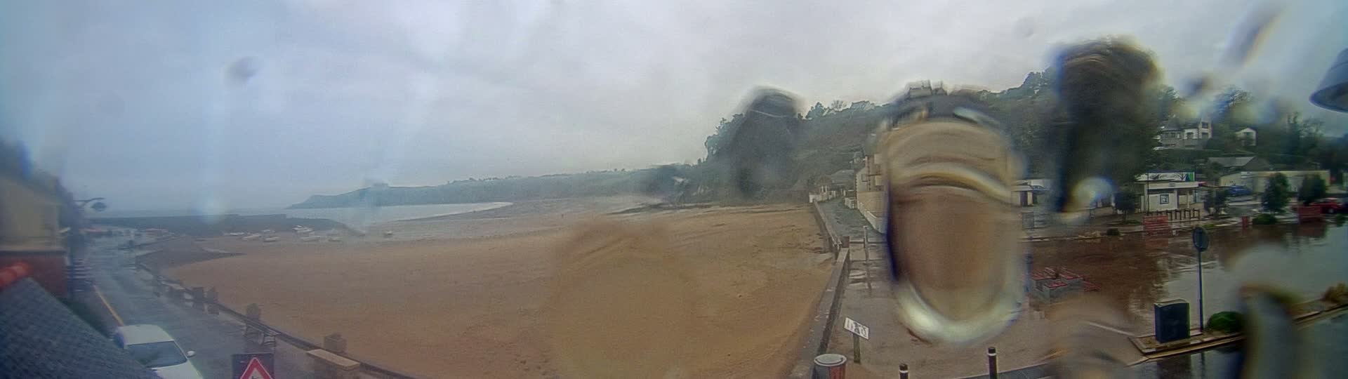 A rain-streaked view shows a sandy beach, a calm body of water, buildings on a hillside, and a wet road in the foreground.