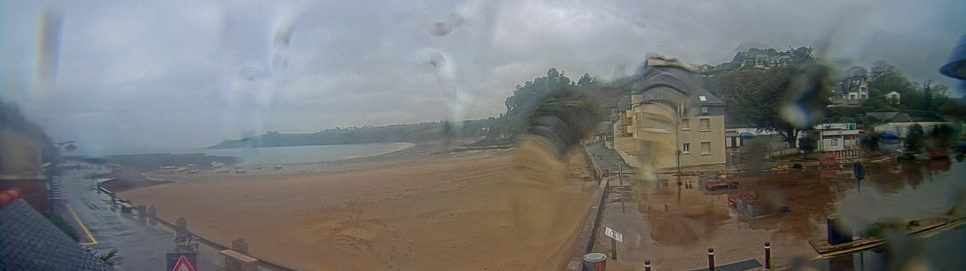 A rain-covered view shows a sandy beach and calm ocean next to buildings and a flooded street.