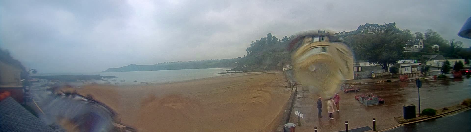 A rain-soaked sandy beach stretches alongside a wet parking lot and buildings, with a calm sea and distant hills visible under a cloudy sky.