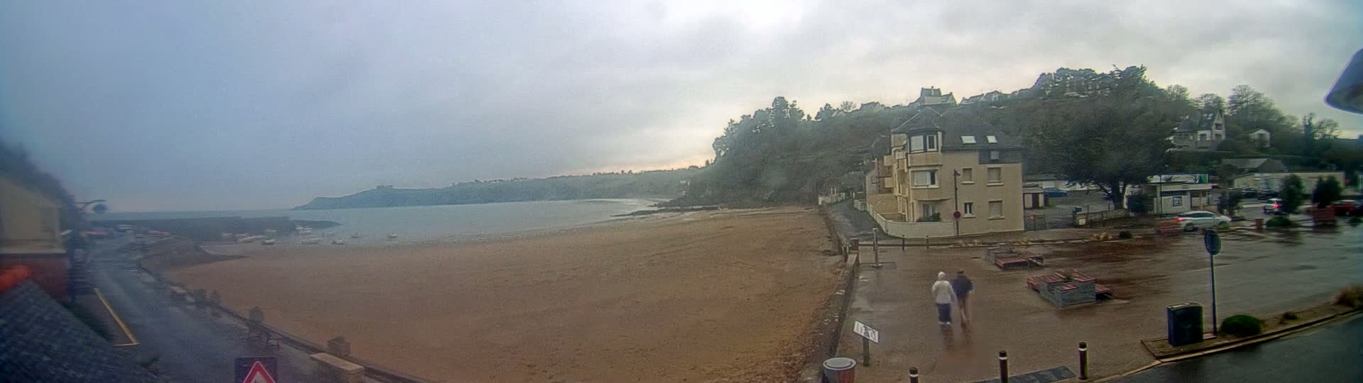 A sandy beach stretches to a calm bay under an overcast sky, with a residential building and wet streets visible in the foreground.