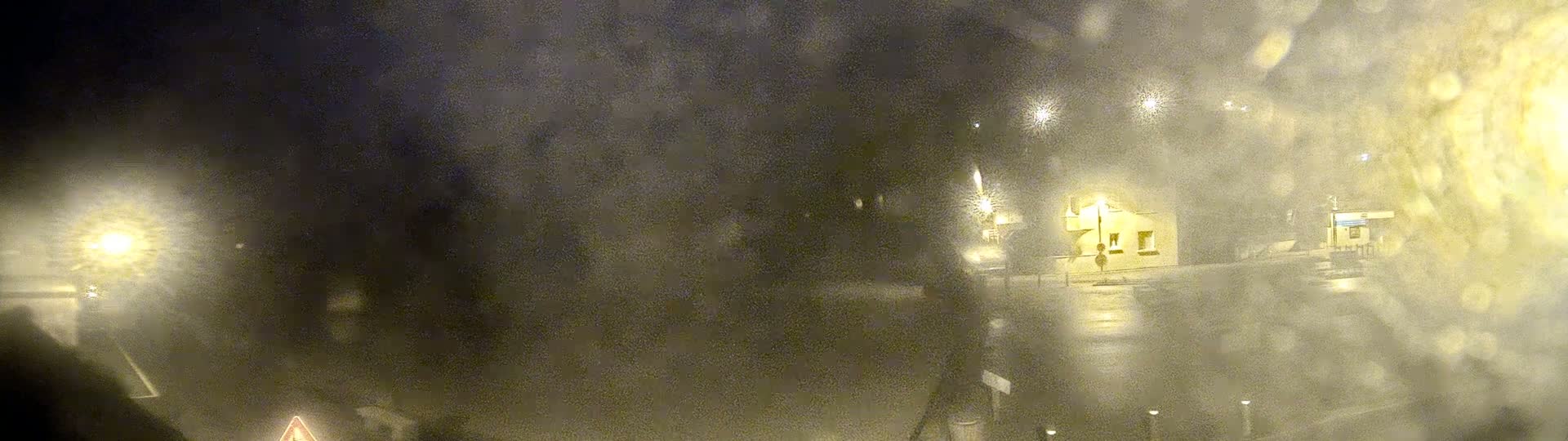 A blurry nighttime view of a street scene, possibly viewed through a rain-covered window, shows dimly lit buildings and streetlights in the distance.