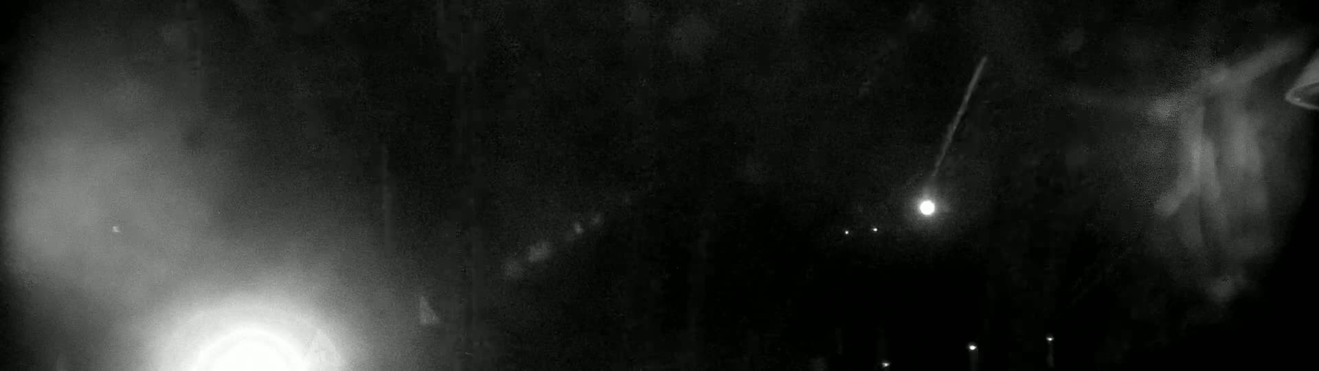 A nighttime image shows a bright streak of light, possibly a meteor, across a dark sky with several faint lights visible.