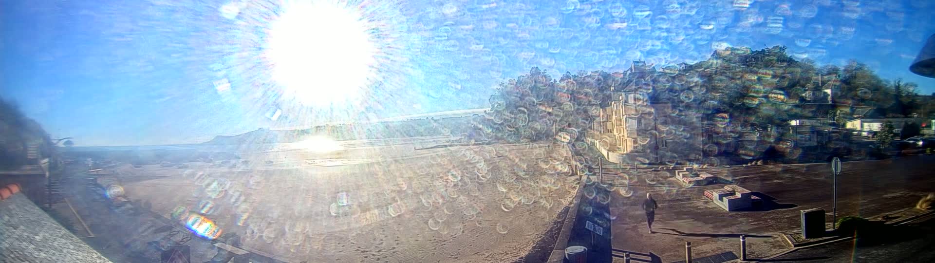 A sunny day reveals a sandy beach, buildings and trees on a hillside, and a person walking along a roadway.