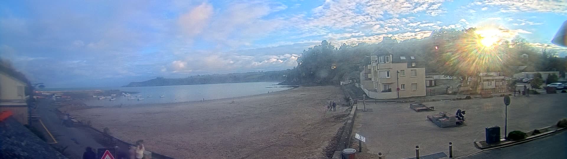 A sandy beach with a calm bay and a few people is visible beneath a mostly sunny sky, with buildings and trees lining the shore in the background.