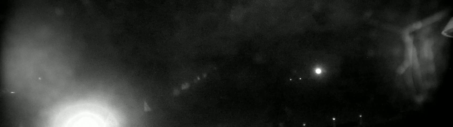 A nighttime, blurry image shows a bright light in the lower-left corner and several fainter lights scattered across a dark, hazy background.